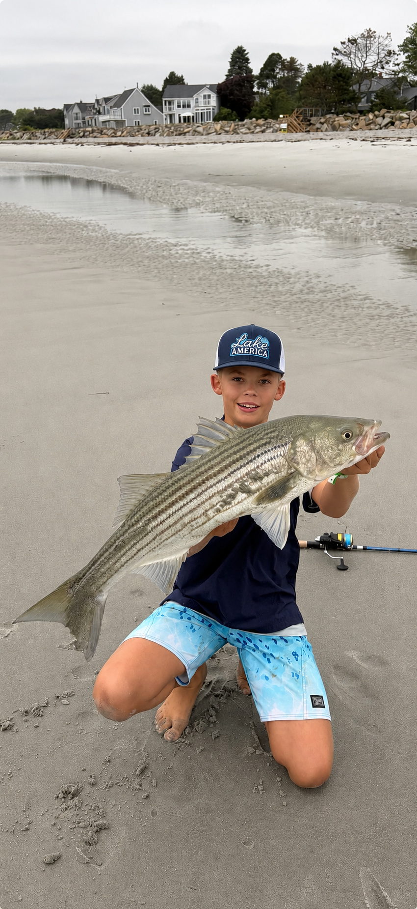 Young angler wearing a Lake America hat while holding a striped bass on the beach